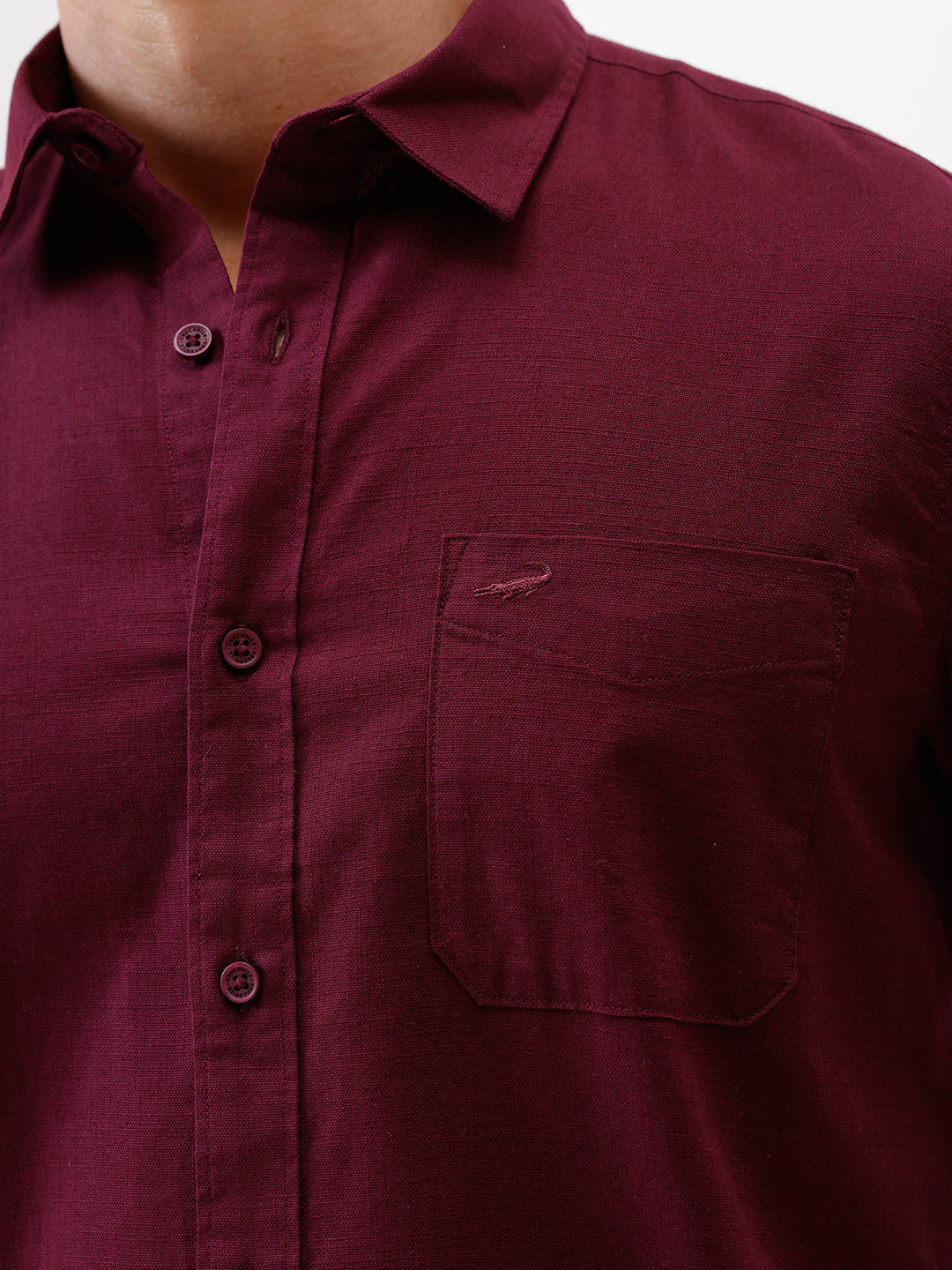 A man wearing a short-sleeve button-up shirt in maroon with a chest pocket, paired with blue jeans, standing against a plain white studio background.