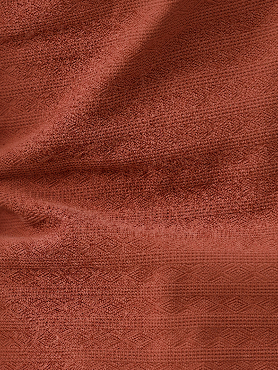 A man wearing a rust-orange short-sleeve polo shirt with a subtle horizontal stripe pattern, paired with dark jeans, standing against a plain white studio background.