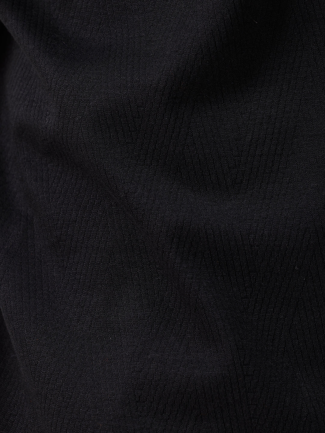 A man wearing a black short-sleeve polo shirt with a single chest pocket, paired with beige trousers, standing against a plain white studio background.