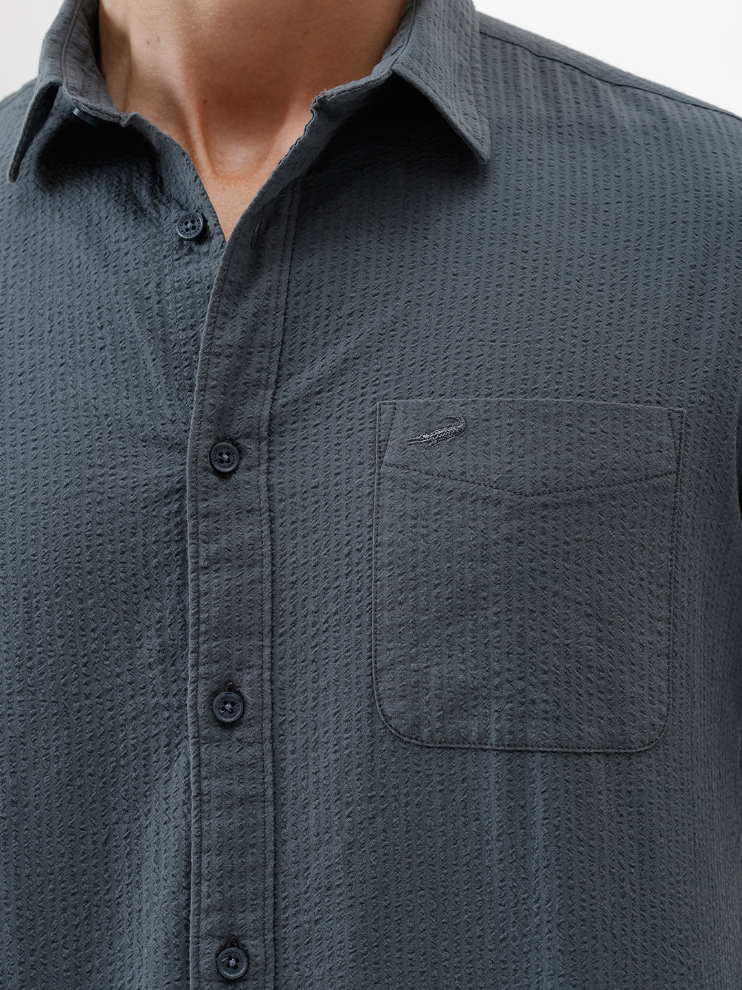 A man wearing a dark charcoal gray long-sleeve button-down shirt with a chest pocket, paired with light gray trousers, standing against a plain white studio background.