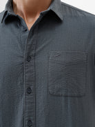 A man wearing a dark charcoal gray long-sleeve button-down shirt with a chest pocket, paired with light gray trousers, standing against a plain white studio background.