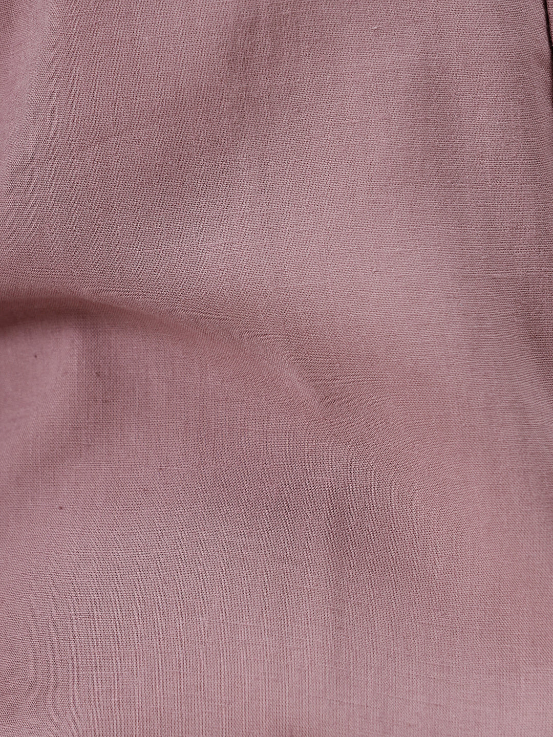 A man wearing a short-sleeve button-up shirt in dusty rose pink with dark blue jeans, standing against a plain white studio background.