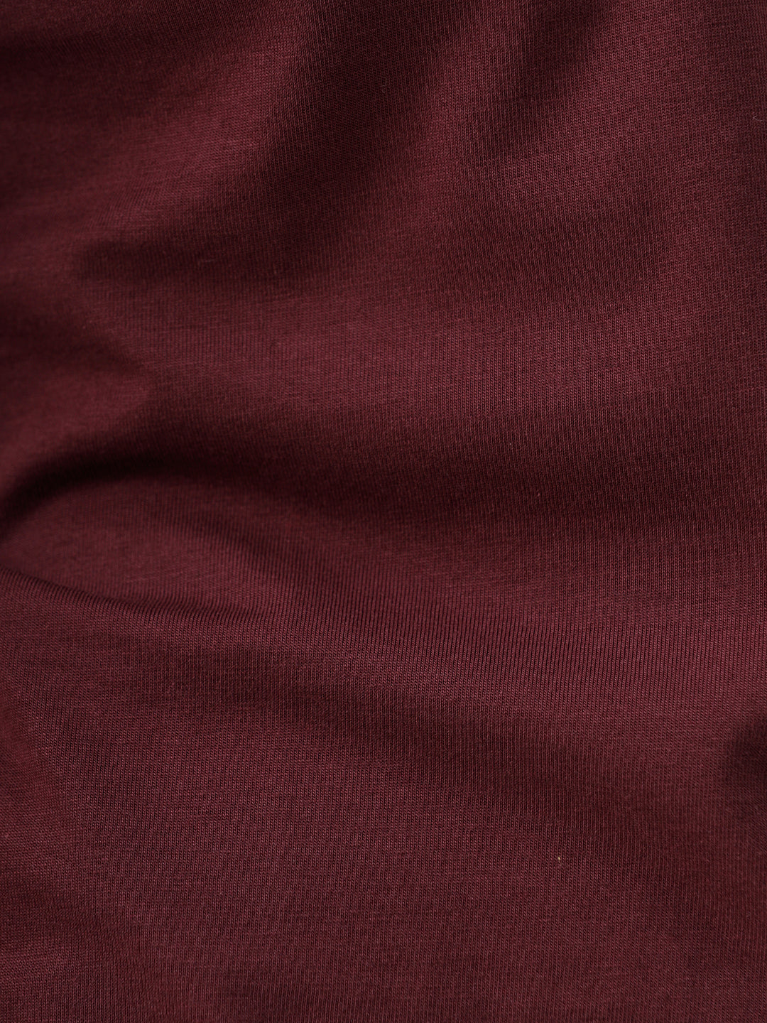 A man wearing a short-sleeve crewneck t-shirt in maroon, paired with dark jeans, standing against a plain light gray studio background.