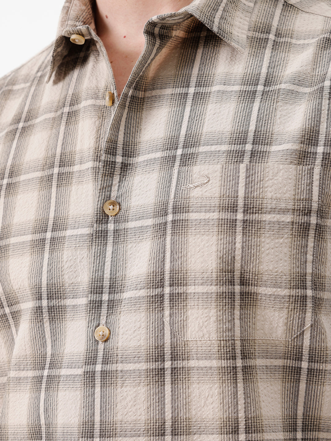 A man wearing a long-sleeve button-up plaid shirt in beige and brown tones, paired with dark jeans, standing against a plain white studio background.