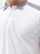 A man modeling a white short-sleeve polo shirt with gray shoulder panels and gray striped trim on the sleeves, paired with gray trousers and black sunglasses, against a plain white background.