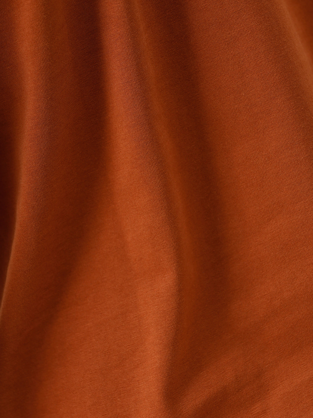 A man wearing a rust-orange crew-neck t-shirt and dark blue jeans, standing against a light gray studio background.