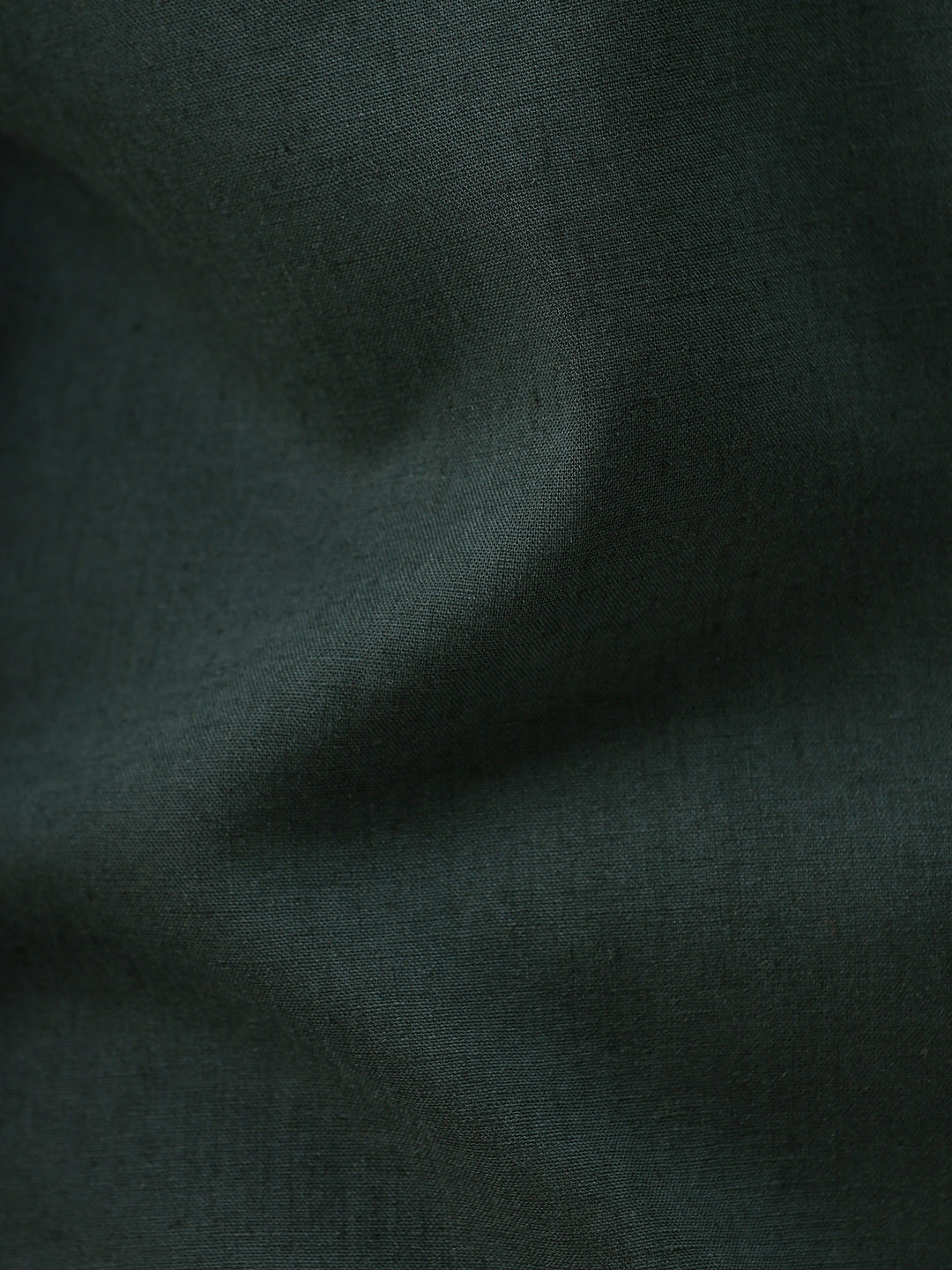 A man wearing a dark forest green short-sleeve button-up shirt with a chest pocket, paired with black jeans, standing against a plain white studio background.