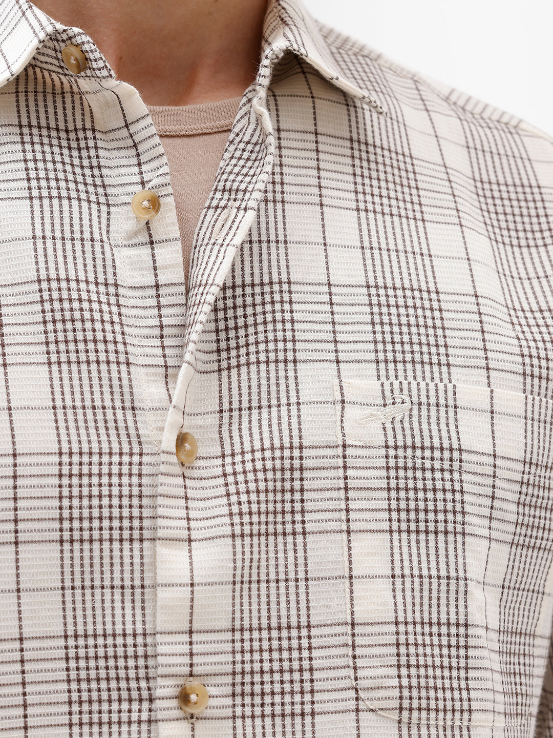A man wearing a long-sleeve button-up plaid shirt in beige, white, and gray, paired with dark pants, standing against a plain white studio background.
