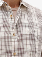 A man wearing a long-sleeve button-up plaid shirt in beige, white, and gray, paired with dark pants, standing against a plain white studio background.