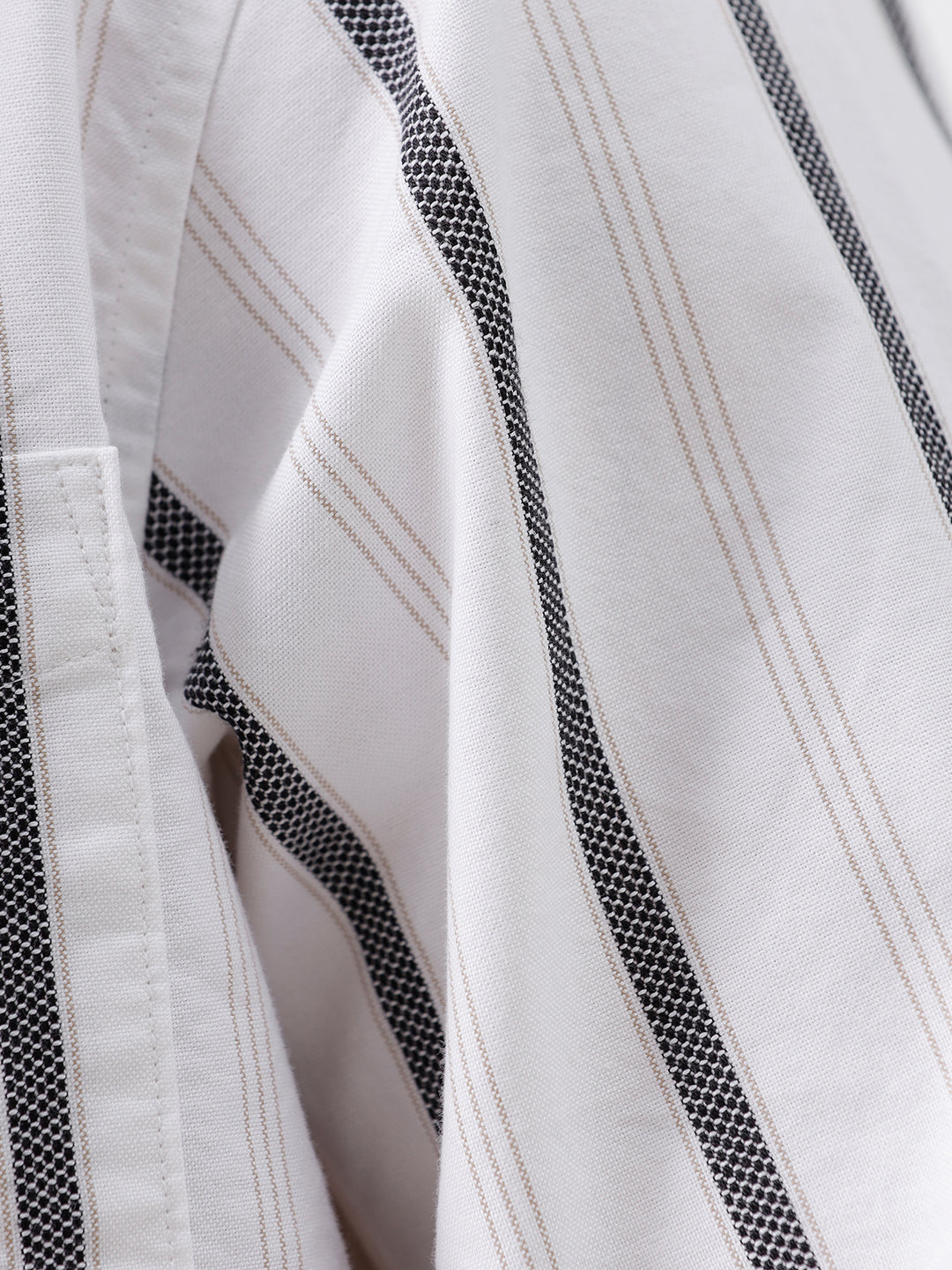 A man wearing a white long-sleeve button-up shirt with vertical gray stripes and beige trousers, standing against a light gray studio background.