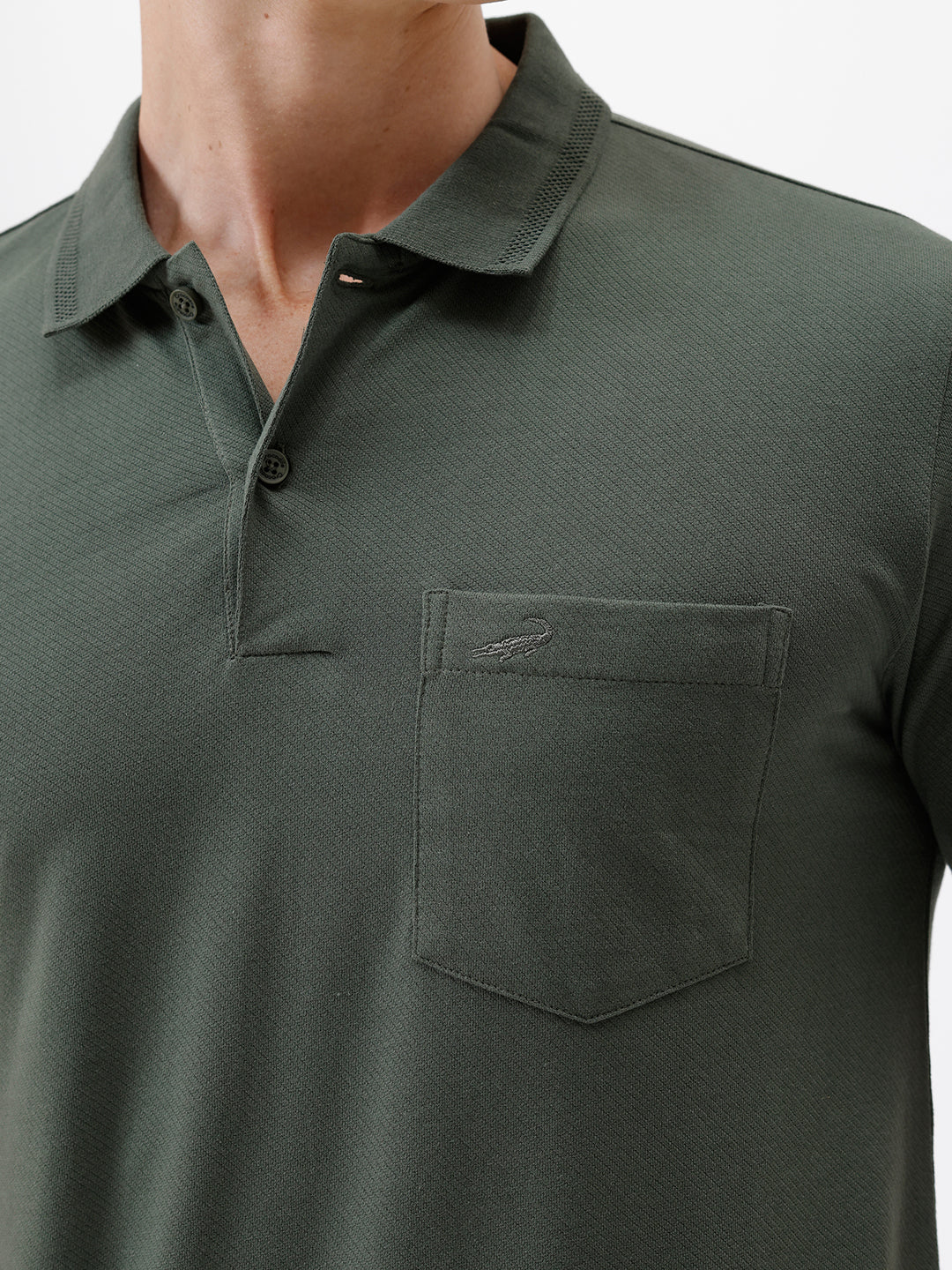 A man wearing a dark olive green short-sleeve polo shirt with a chest pocket, paired with dark jeans, standing against a plain light gray background.