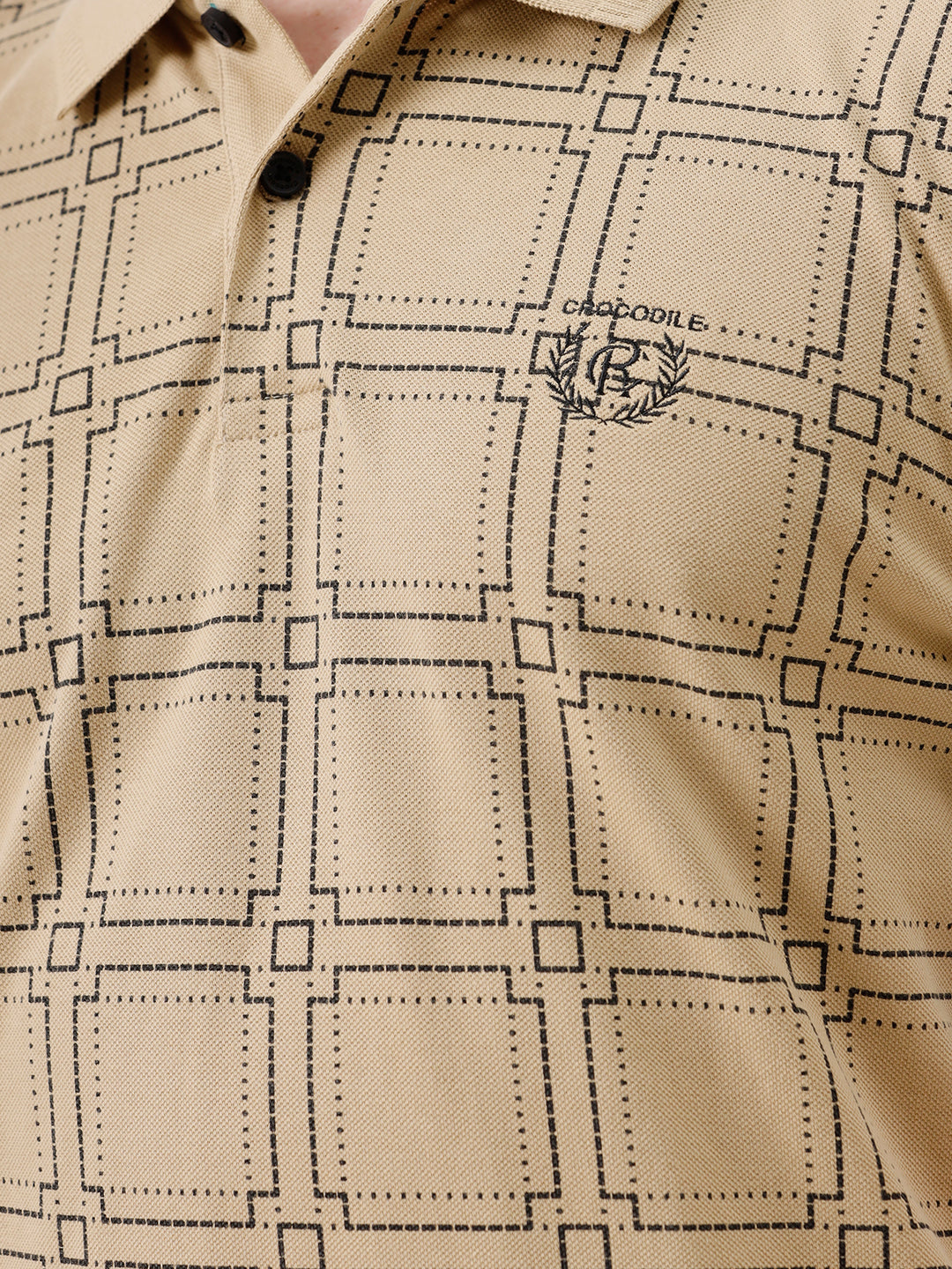A man wearing a beige short-sleeve polo shirt with a brown grid pattern, paired with black jeans, standing against a plain white studio background.