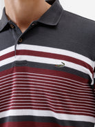A man wearing a dark gray Lacoste polo shirt with horizontal red and white stripes across the chest and sleeves, paired with blue jeans, against a plain light gray background.