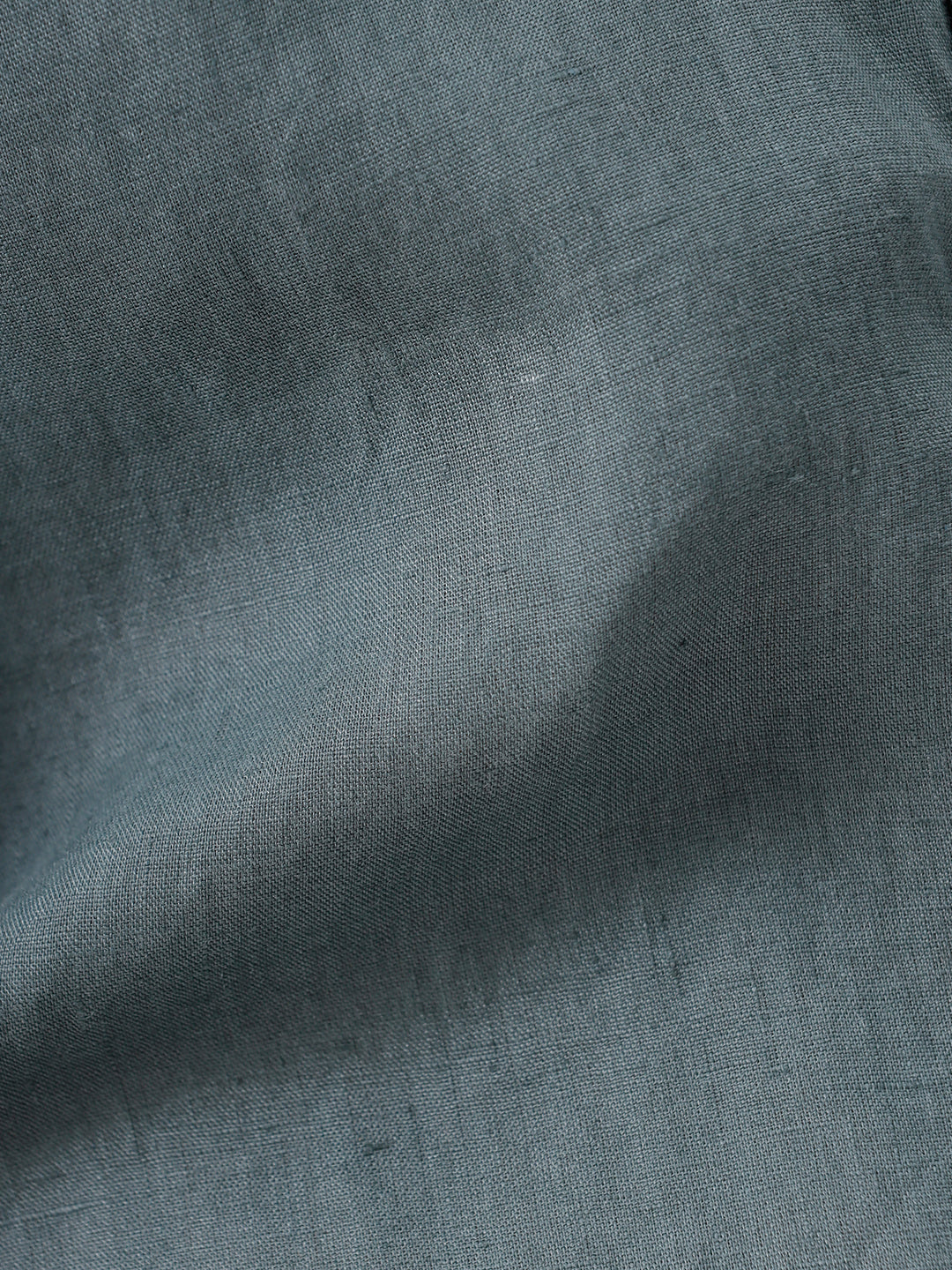 A man wearing a short-sleeve button-up shirt in a muted slate green color, paired with light beige trousers, standing against a plain light gray background.