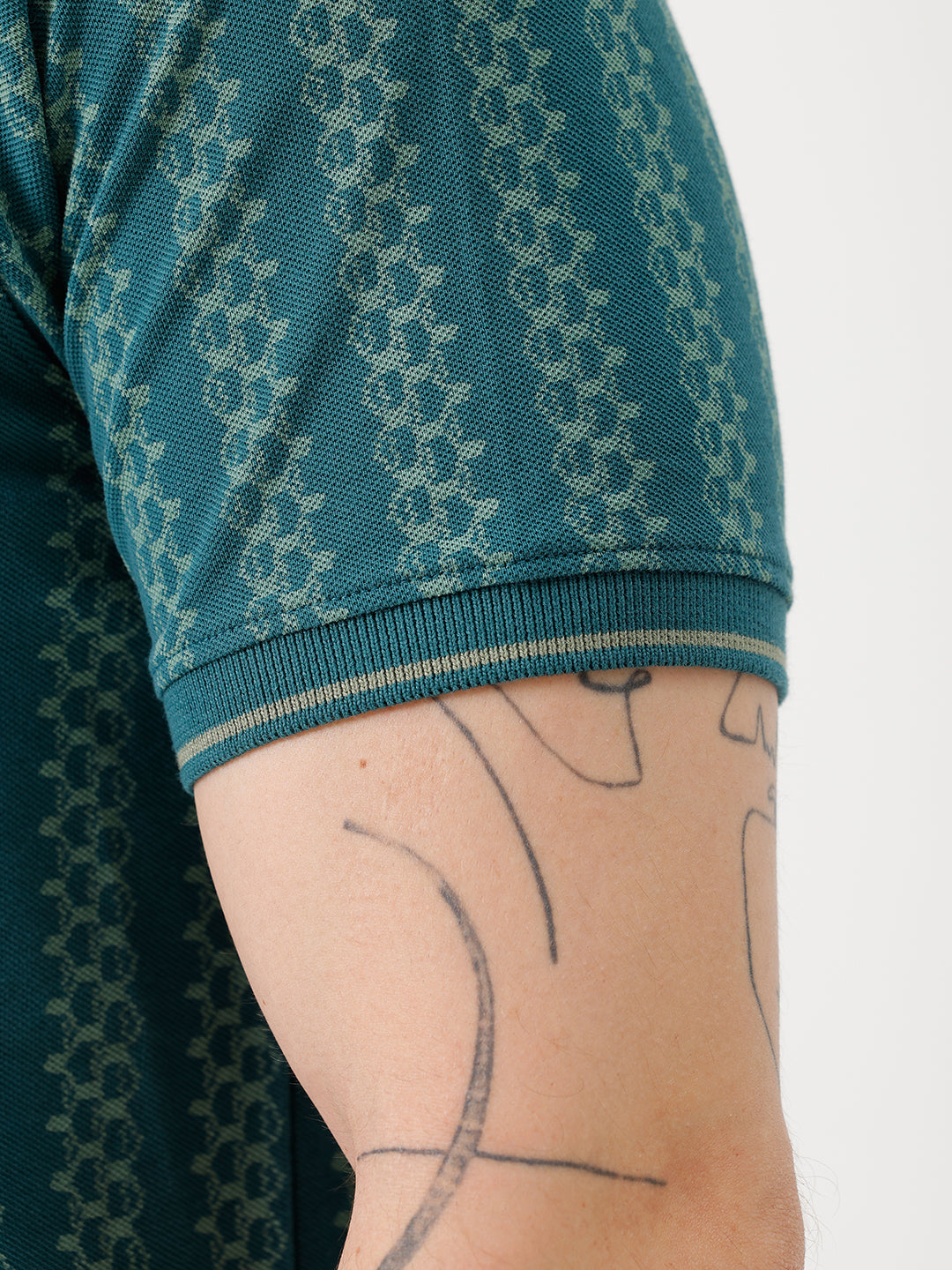 A man wearing a teal green short-sleeve polo shirt with a white geometric pattern, paired with black jeans, standing against a plain white studio background.