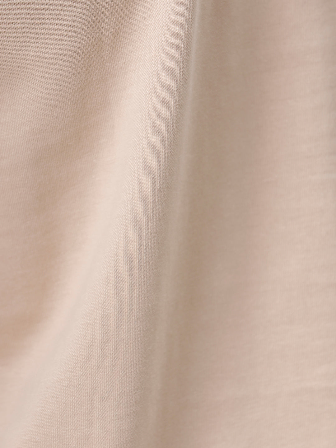 A man wearing a beige short-sleeve crewneck t-shirt with a subtle tonal logo on the left chest, paired with black jeans, standing against a light gray studio background.