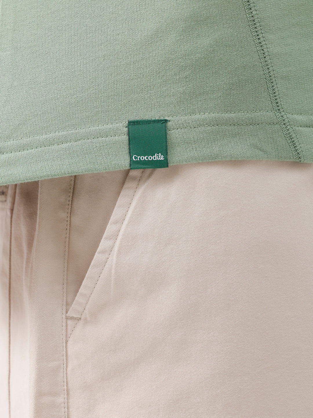 A man wearing a light sage green athletic t-shirt with a small tonal logo on the chest, paired with beige pants, standing against a plain white studio background.
