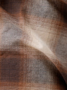 A man wearing a long-sleeve button-up plaid shirt in brown, beige, and gray tones, paired with dark jeans, standing against a plain white studio background.