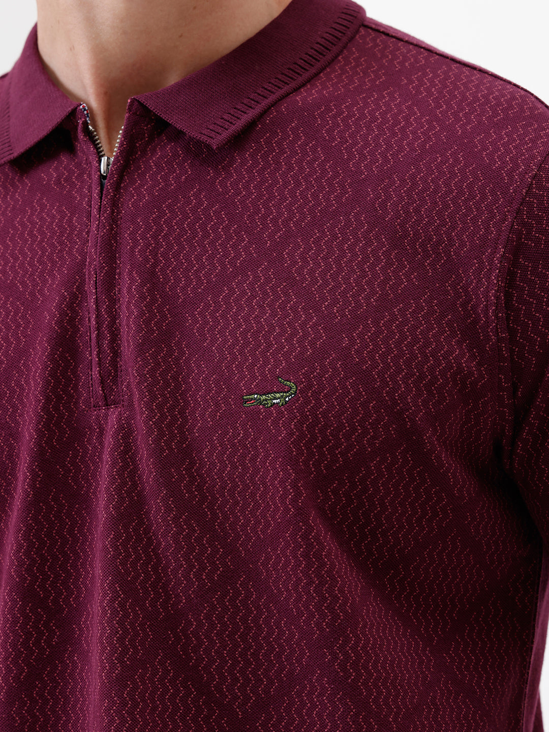 A man wearing a short-sleeve polo shirt in deep burgundy with a subtle textured geometric pattern, styled with gray trousers, sunglasses, and a watch against a plain white background.