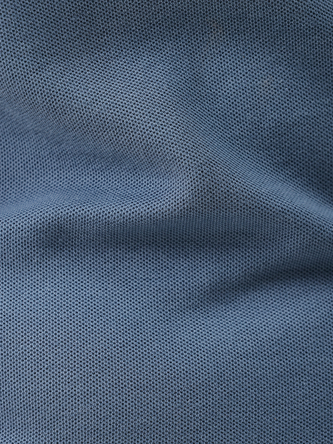 A man wearing a slate blue short-sleeve polo shirt and beige trousers, standing against a plain white studio background.