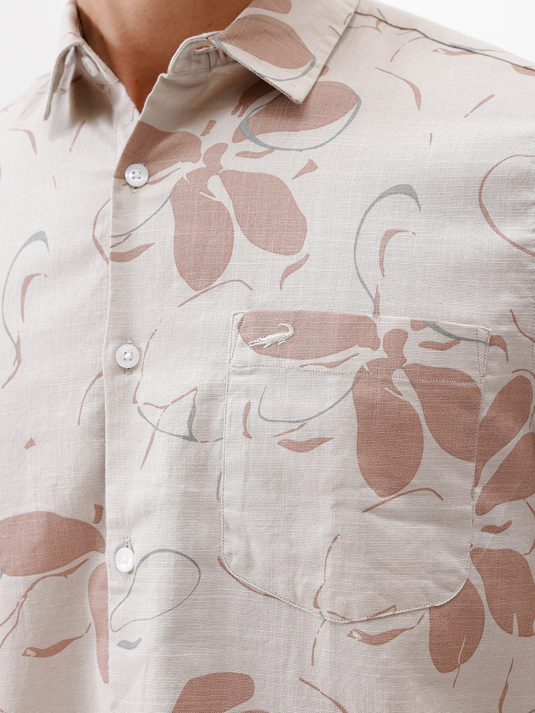 A man wearing a short-sleeve button-up shirt with a beige and brown floral pattern, paired with gray trousers, standing against a plain white background.