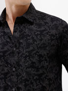 A man wearing a black long-sleeve button-up shirt with a subtle tonal floral pattern, paired with beige cargo pants and dark sunglasses, standing against a plain white background.