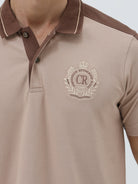 A man modeling a short-sleeve polo shirt in beige with brown shoulder panels and collar trim, paired with dark blue jeans, against a light gray studio background.