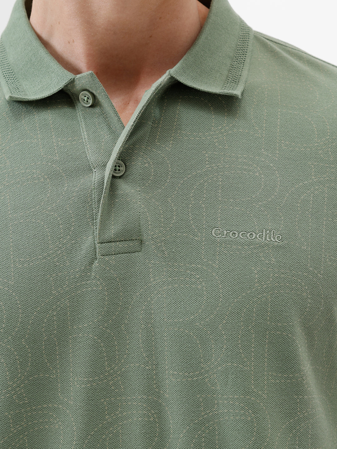 A man wearing a short-sleeve polo shirt in sage green, paired with dark blue jeans, standing against a plain light gray background.