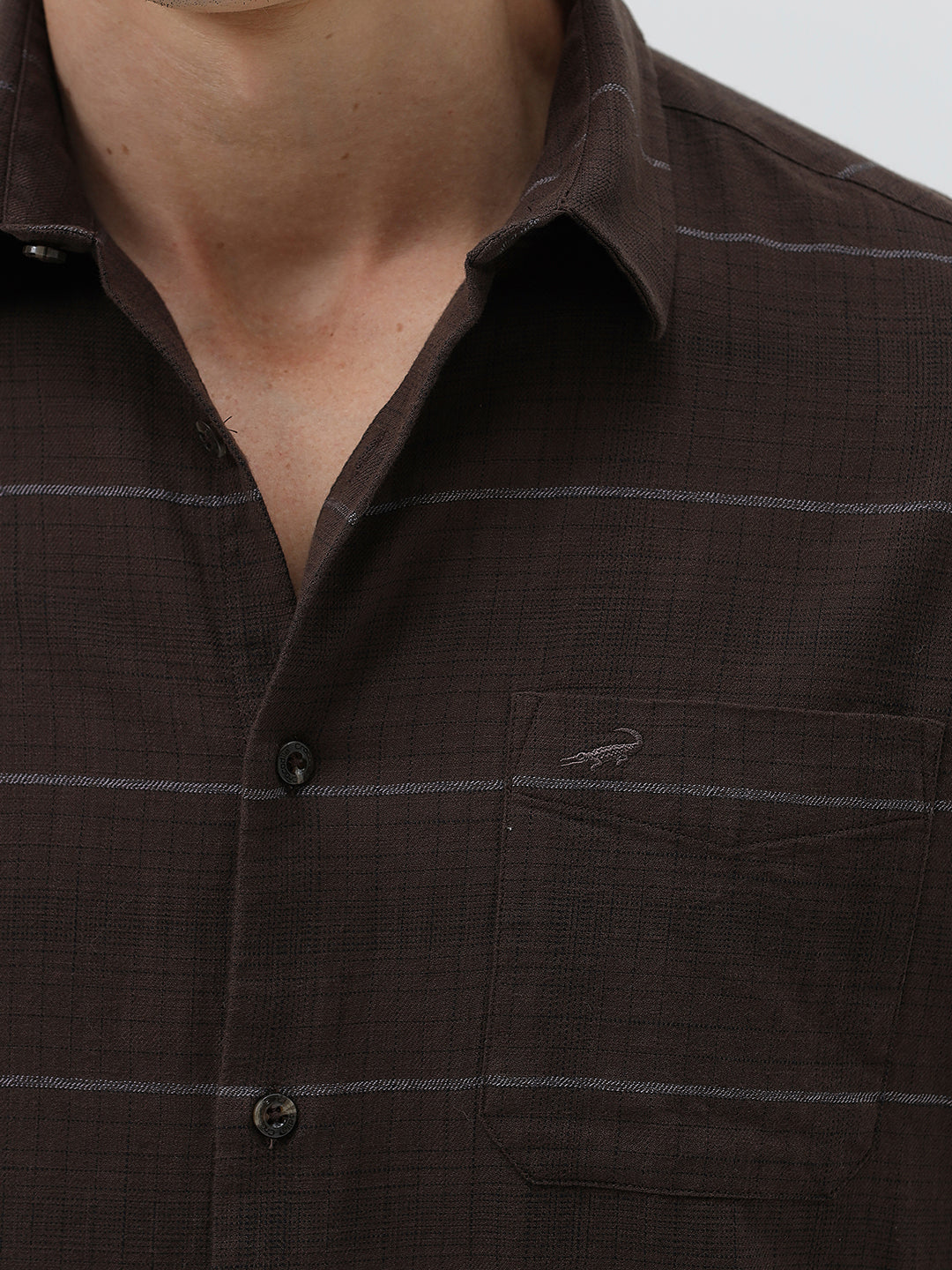 A man wearing a dark brown long-sleeve button-up shirt with thin horizontal stripes, paired with dark blue jeans, standing against a light gray studio background.