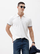 A man wearing a white short-sleeve polo shirt with gray ribbed trim on the collar and sleeve cuffs, paired with dark blue jeans, photographed against a plain white studio background.