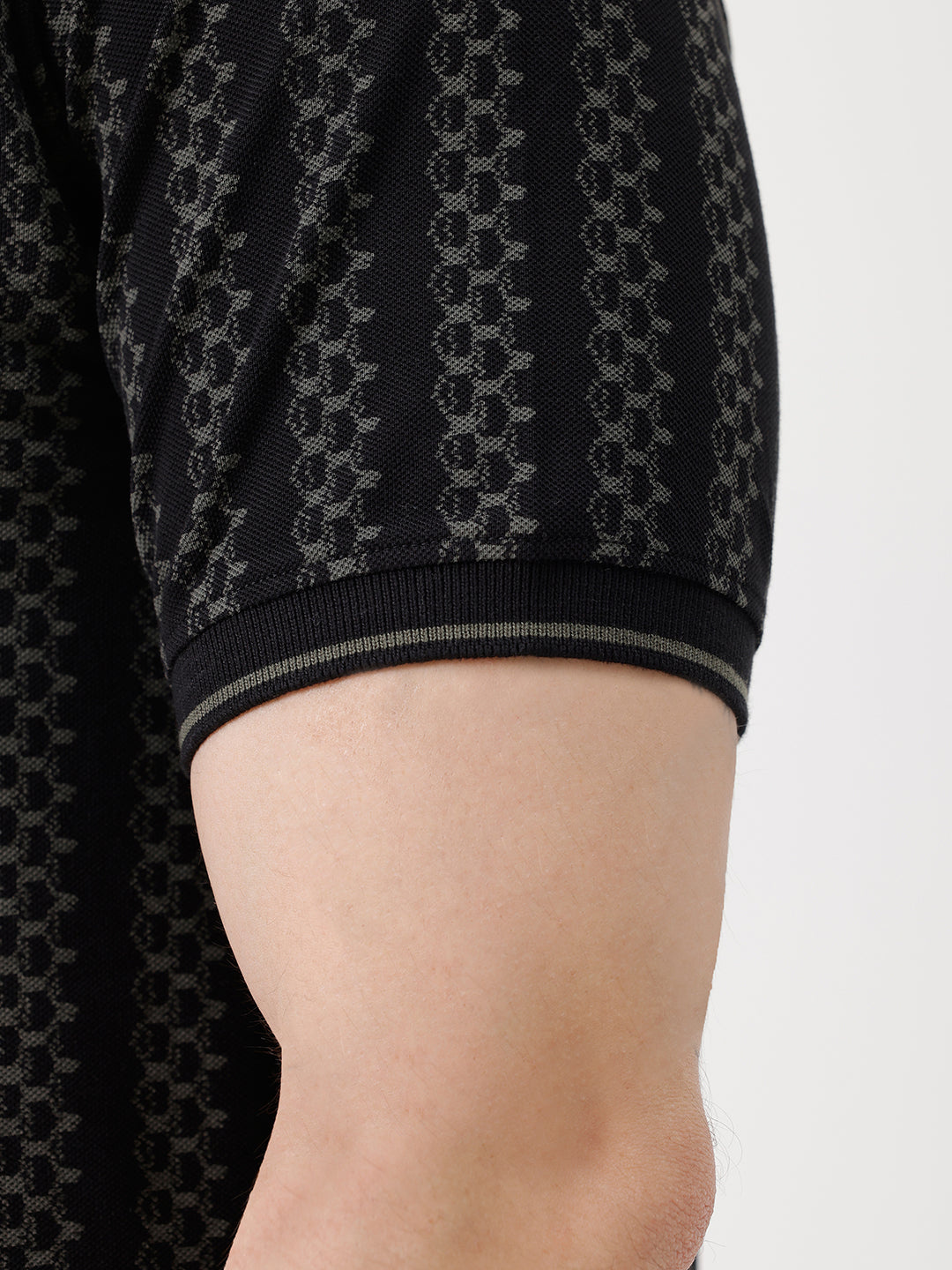 A man wearing a black short-sleeve polo shirt with a subtle geometric pattern, paired with dark gray trousers, standing against a plain white studio background.