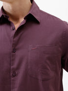 A man modeling a short-sleeve button-up shirt in a deep plum purple color, paired with gray trousers, against a plain white studio background.