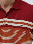 A man wearing a red short-sleeve polo shirt with horizontal cream and rust-orange stripes across the chest and sleeves, paired with dark blue jeans and black sunglasses, against a light gray studio background.