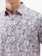 A man wearing a short-sleeve button-up shirt with a gray and white abstract pattern, paired with dark gray trousers, standing against a plain white studio background.