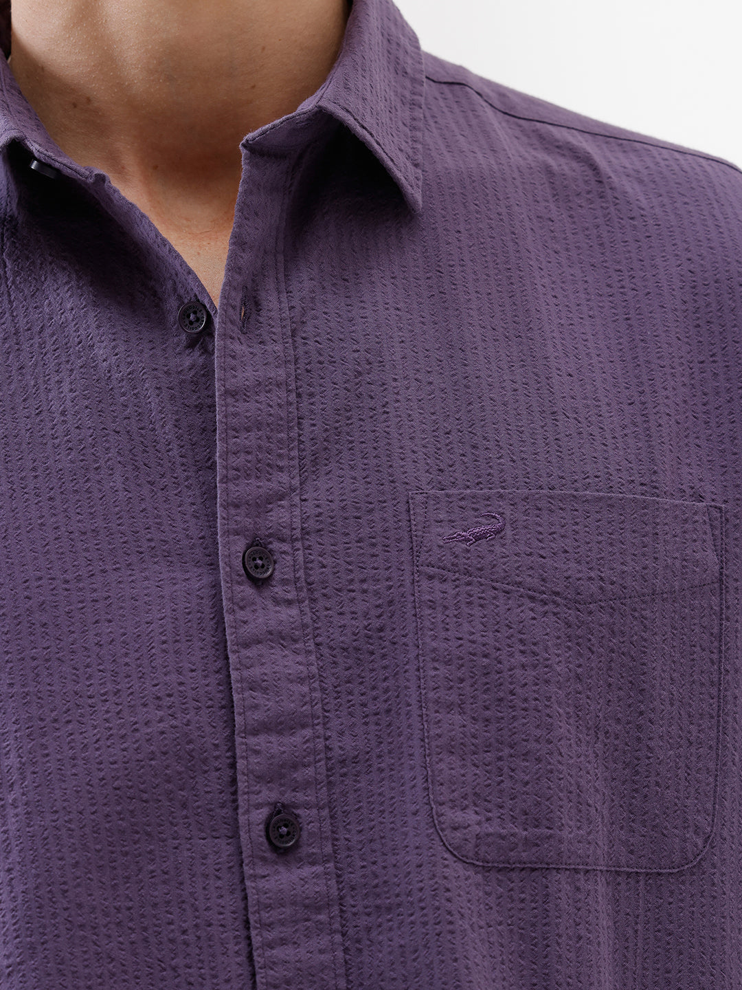 A man wearing a long-sleeve button-down shirt in a muted purple color, paired with black pants, standing against a plain white studio background.