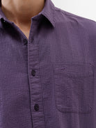 A man wearing a long-sleeve button-down shirt in a muted purple color, paired with black pants, standing against a plain white studio background.