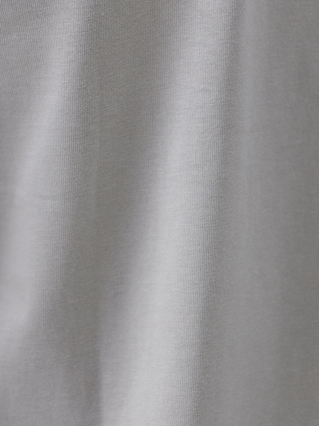 A man wearing a light gray short-sleeve crewneck t-shirt with a subtle tonal chest logo, paired with dark jeans, standing against a light gray studio background.