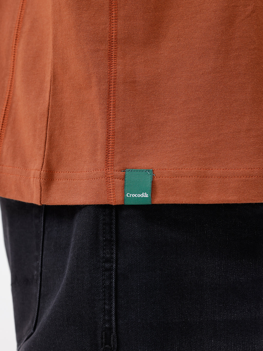 A man wearing a rust-orange crewneck t-shirt with a subtle tonal 'ELECTRIC' logo across the chest, paired with black pants, photographed against a clean white studio background.