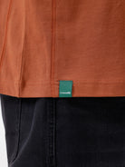 A man wearing a rust-orange crewneck t-shirt with a subtle tonal 'ELECTRIC' logo across the chest, paired with black pants, photographed against a clean white studio background.
