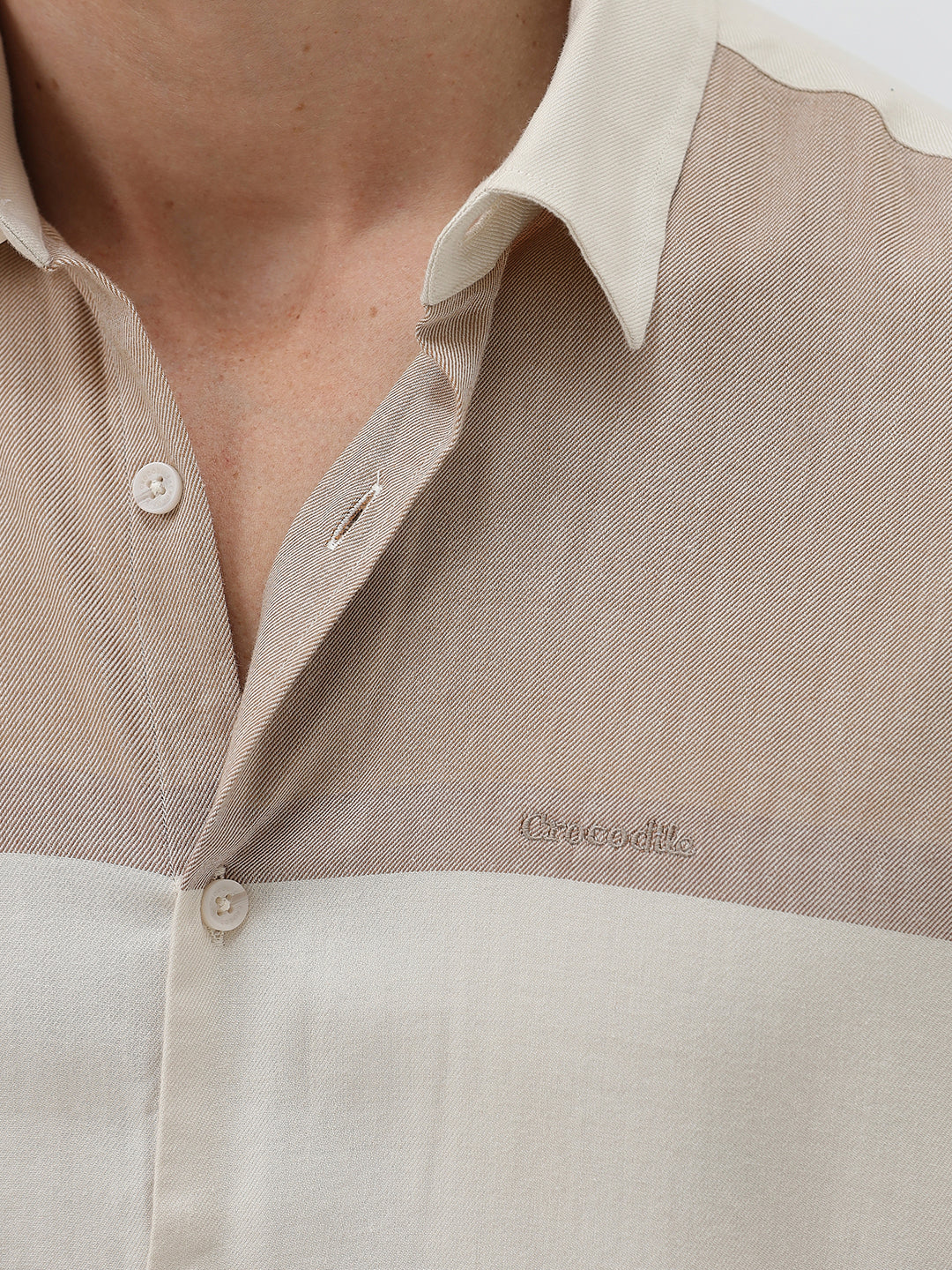 A man wearing a short-sleeve linen button-up shirt with a color-blocked design in beige and cream, paired with black pants, against a plain white studio background.