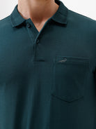 A man wearing a dark teal short-sleeve polo shirt with a chest pocket, paired with beige cargo pants, standing against a plain white studio background.