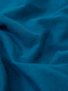A man wearing a teal blue short-sleeve t-shirt with a subtle tonal 'GREATNESS' text graphic across the chest, paired with black pants, photographed against a clean white studio background.