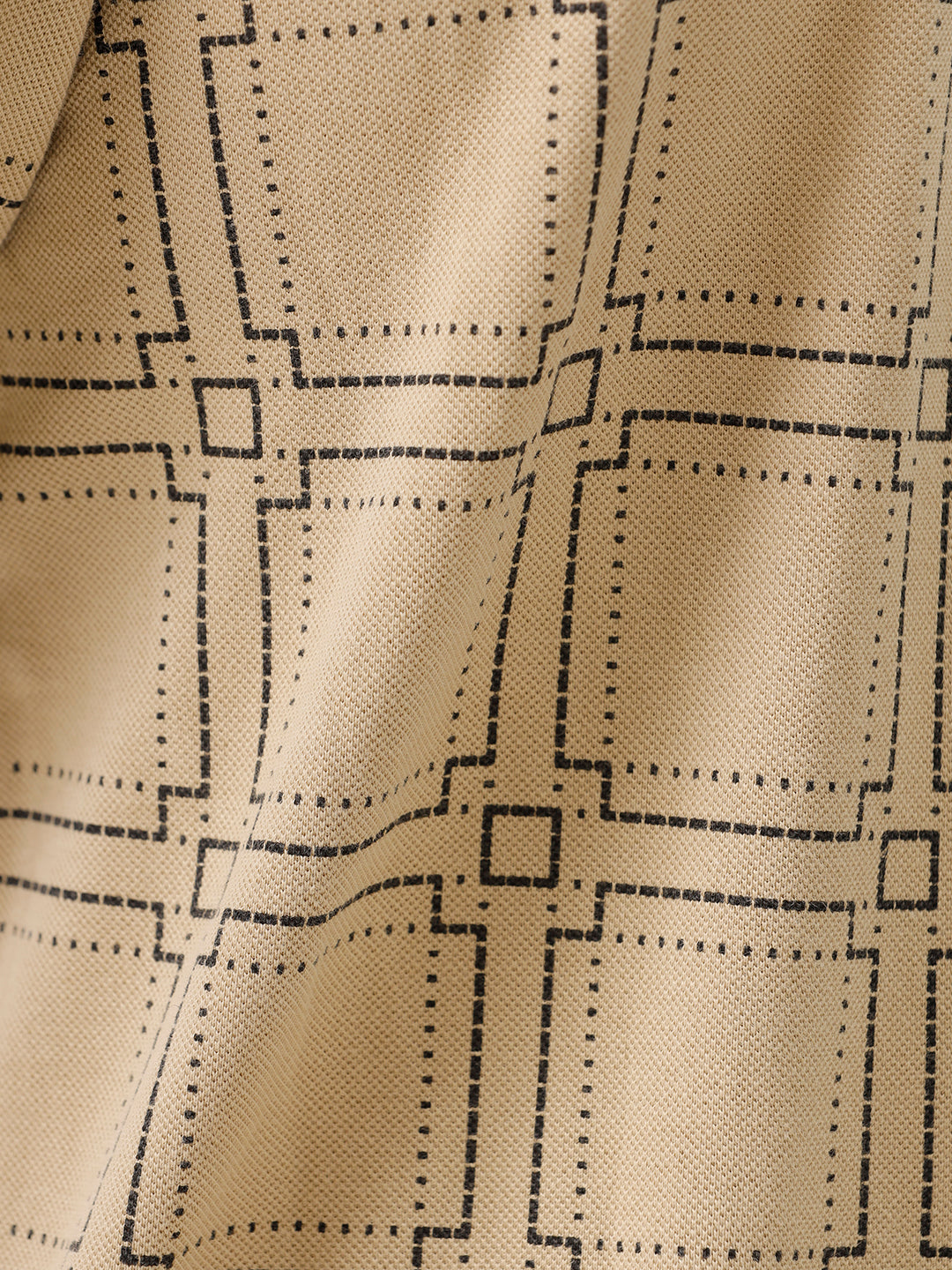 A man wearing a beige short-sleeve polo shirt with a brown grid pattern, paired with black jeans, standing against a plain white studio background.