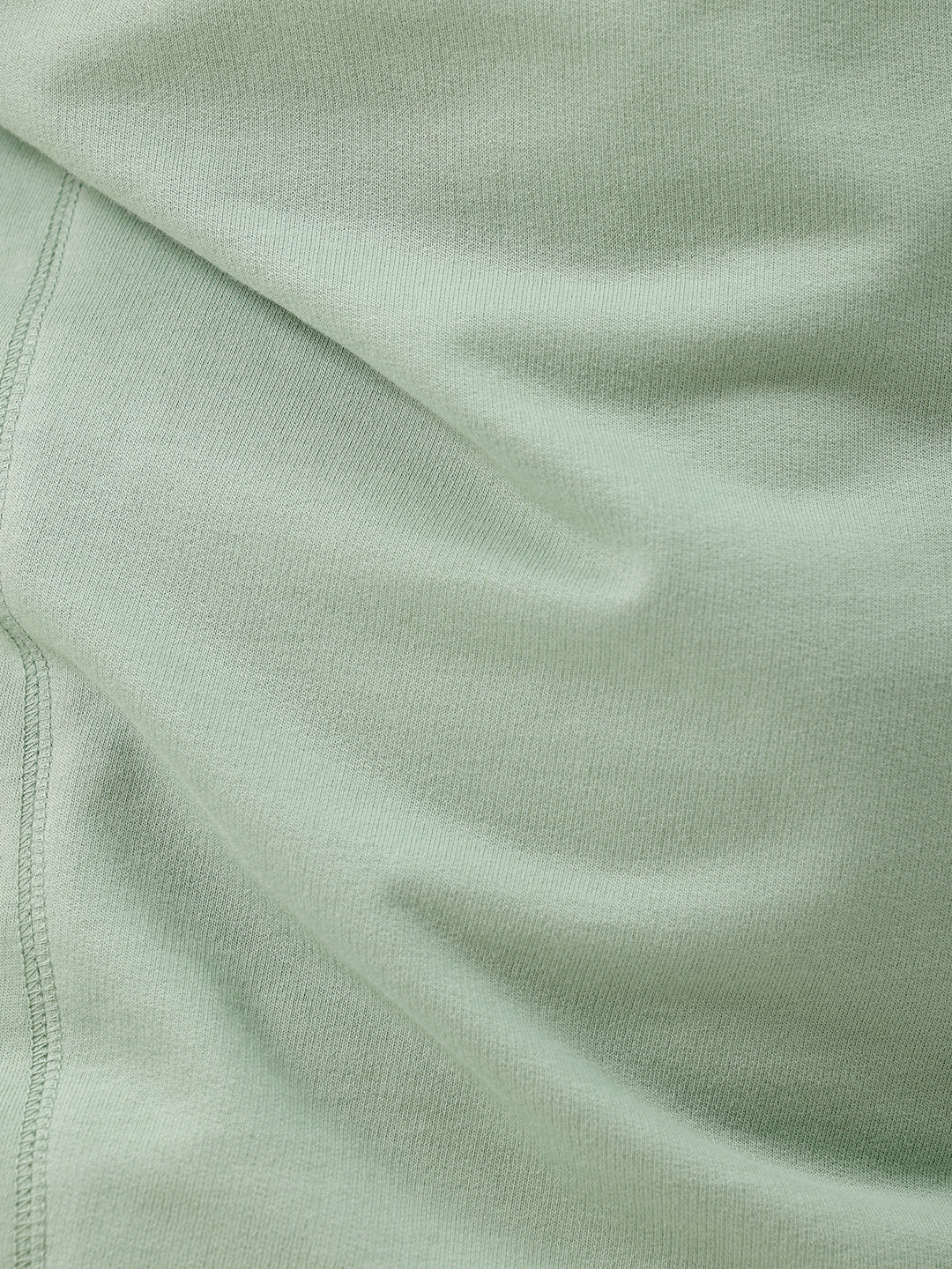 A man wearing a light sage green athletic t-shirt with a small tonal logo on the chest, paired with beige pants, standing against a plain white studio background.