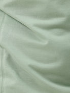 A man wearing a light sage green athletic t-shirt with a small tonal logo on the chest, paired with beige pants, standing against a plain white studio background.