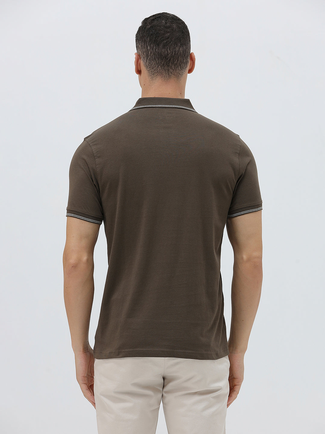 A man wearing a dark brown short-sleeve polo shirt with contrasting light gray trim on the collar and sleeve cuffs, paired with off-white trousers, against a plain light gray background.