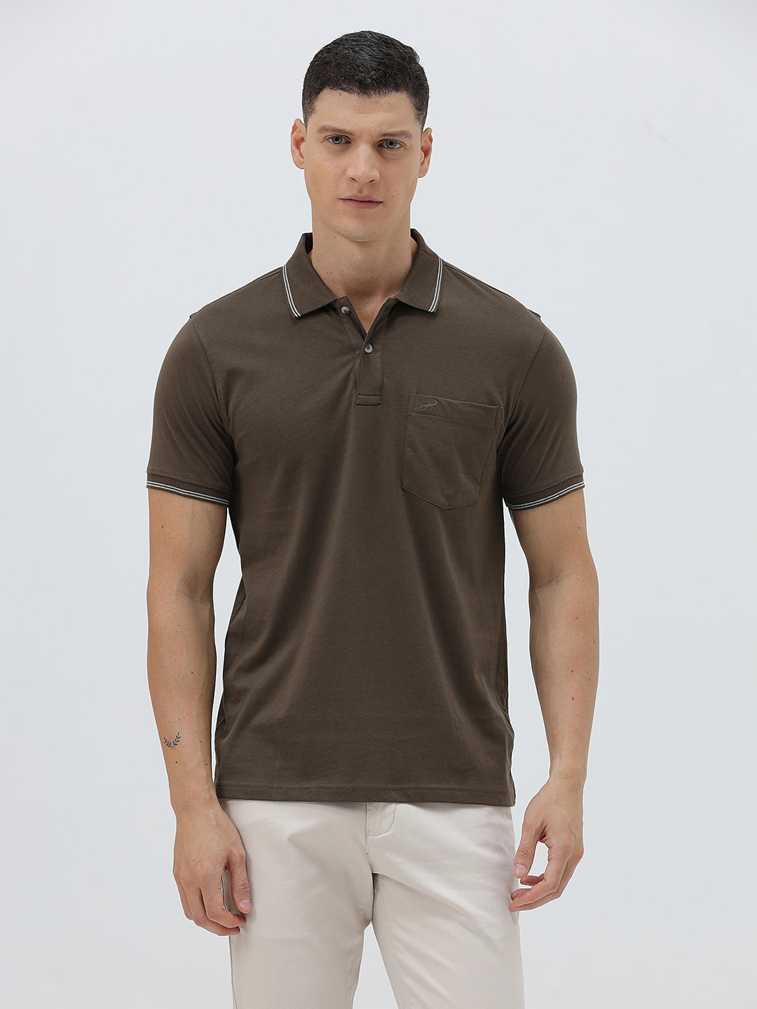 A man wearing a dark brown short-sleeve polo shirt with contrasting light gray trim on the collar and sleeve cuffs, paired with off-white trousers, against a plain light gray background.