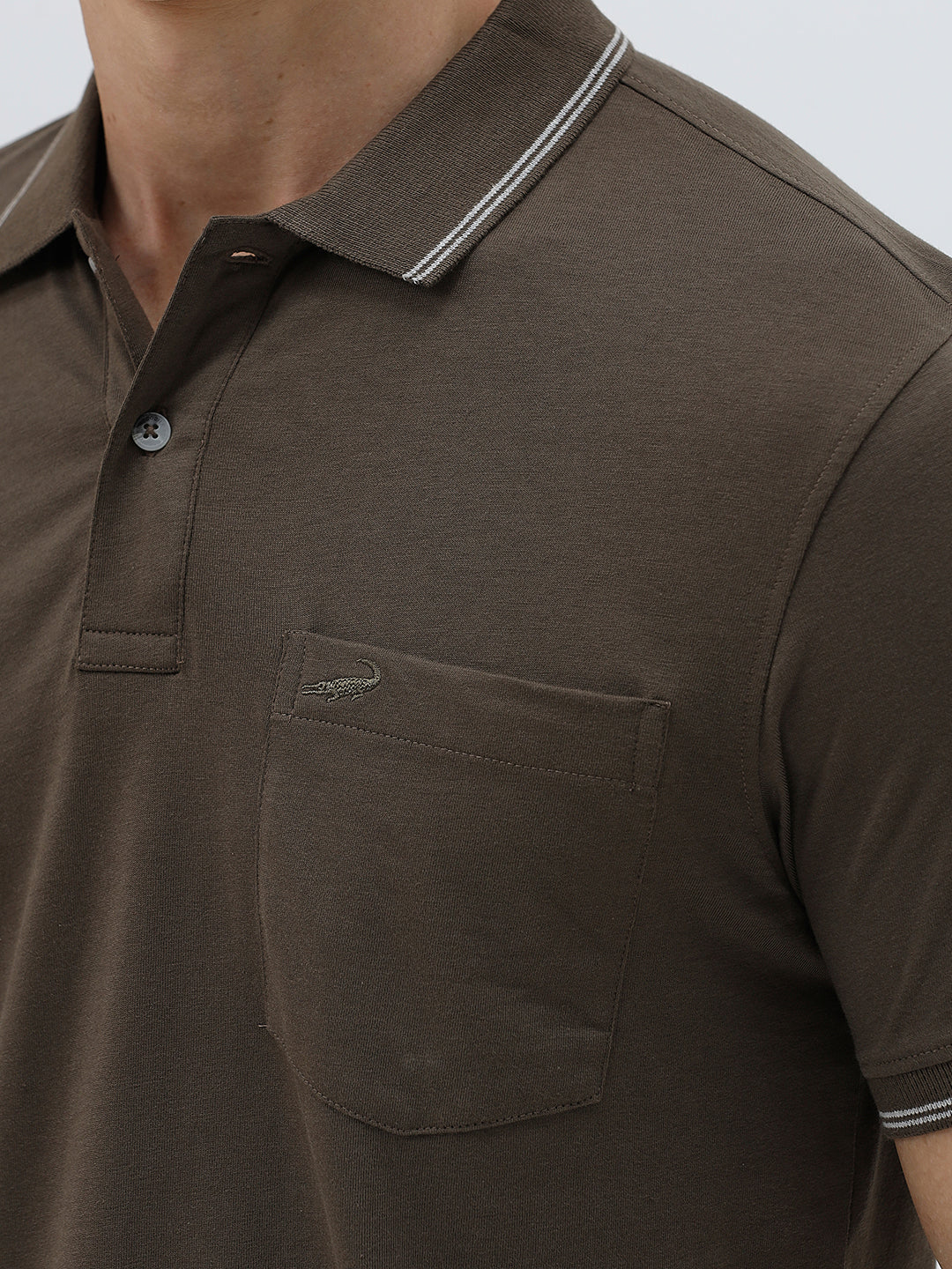 A man wearing a dark brown short-sleeve polo shirt with contrasting light gray trim on the collar and sleeve cuffs, paired with off-white trousers, against a plain light gray background.
