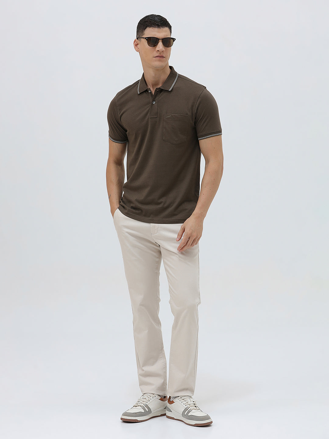 A man wearing a dark brown short-sleeve polo shirt with contrasting light gray trim on the collar and sleeve cuffs, paired with off-white trousers, against a plain light gray background.