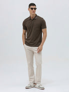 A man wearing a dark brown short-sleeve polo shirt with contrasting light gray trim on the collar and sleeve cuffs, paired with off-white trousers, against a plain light gray background.